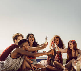 Eine Gruppe von Freunden sitzt im Freien und stößt mit Getränken an. Fröhliche junge Leute trinken ein Bier an einem Sommertag. - JLPSF23528