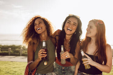 Cheerful girls in casuals having fun outdoors. Diverse group of three young women walking outdoors with beers and laughing. - JLPSF23534