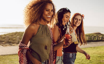 Hübsche Mädchen in Freizeitkleidung, die mit einem Bier im Freien spazieren gehen. Eine Gruppe von drei Frauen, die an einem Sommerwochenende abhängen. - JLPSF23536
