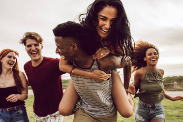Smiling group of friends enjoying on weekend. Multi-ethnic men and women having fun outdoors. Man carrying woman in his back and running with friends trying to chat him. - JLPSF23546