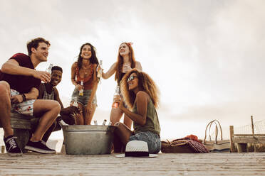 Eine Gruppe von Männern und Frauen sitzt am Strand und trinkt Bier. Fünf junge Freunde feiern am Strand. - JLPSF23551