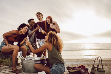 Lächelnde junge Frau mit Bierflasche und Freunden, die am Strand stehen. Eine Gruppe von Freunden feiert abends am Strand - JLPSF23552