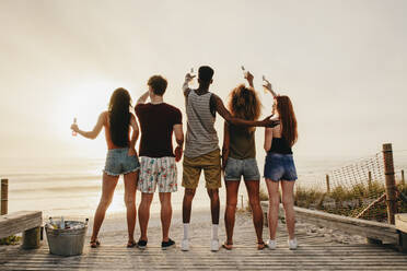 Rückansicht von jungen Freunden, die an der Strandpromenade stehen und eine Bierflasche ins Meer halten. - JLPSF23557