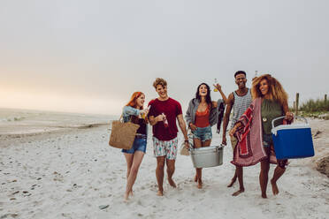 Eine Gruppe junger Leute geht am Strand spazieren und trägt eine Kühlbox und einen Getränkebecher. Junge Männer und eine Frau am Meeresufer. - JLPSF23561