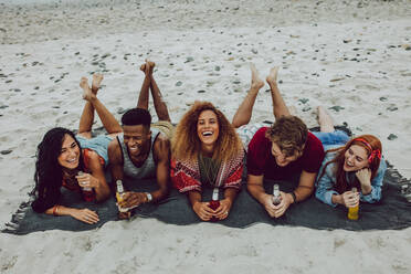 Cheerful young friends lying outdoors on a plaid with beer bottles. Young men and women laughing at the sea shore with beers. - JLPSF23562