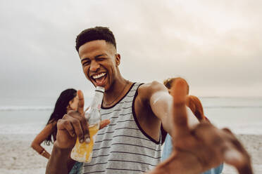 Aufgeregter junger afrikanischer Mann mit Bier am Strand mit Freunden im Hintergrund. Lächelnder Mann mit einer Gruppe von Freunden im Hintergrund am Strand. - JLPSF23567