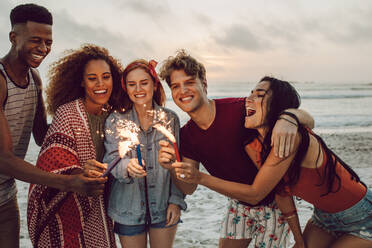 Glückliche Freunde feiern am Strand mit Wunderkerzen. Multi-ethnische Gruppe von Freunden zusammen am Strand feiern mit Feuerwerk. - JLPSF23569