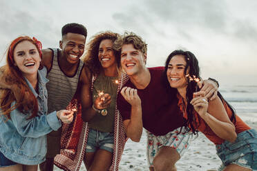 Eine bunte Gruppe junger Leute feiert den Neujahrstag am Strand. Junge Männer und Frauen haben Spaß mit Wunderkerzen im Freien am Meeresufer. - JLPSF23571