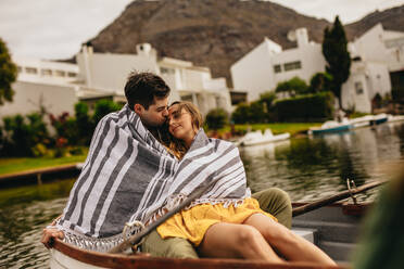 Mann küsst seine Freundin in einem Boot sitzend bei einem Date auf einem See. Junges Paar in ein Handtuch eingewickelt in einem Boot sitzend, das entspannt Zeit miteinander bei einem Date verbringt. - JLPSF23576