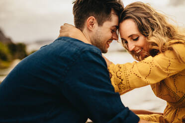 Side view of a young couple sitting together touching their heads. Romantic couple on a date sitting near a lake holding each other. - JLPSF23580