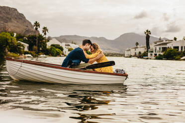 Junges Paar in einem Boot sitzend, die Köpfe berührend, sich gegenseitig haltend. Paar auf einem Bootstermin in einem See mit Hügeln und Häusern im Hintergrund. - JLPSF23581