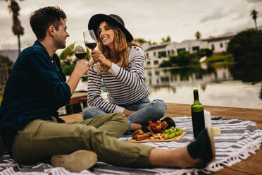 Junges Paar beim Rendezvous stößt mit einem Glas Wein an einem See an. Junge Frau mit Hut sitzt mit ihrem Freund zusammen und trinkt Wein mit Snacks davor. - JLPSF23588