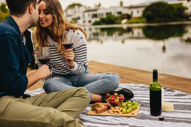 Verliebtes Paar sitzt auf einem Holzsteg in der Nähe eines Sees und trinkt Wein und Snacks. - JLPSF23589