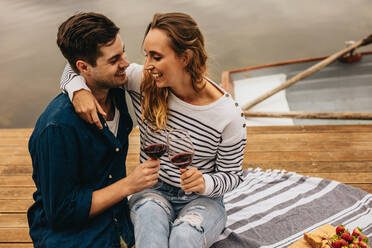 Happy couple drinking wine sitting near a lake and laughing holding each other. Couple in love sitting on a wooden dock beside a lake with wine glasses in hand and snacks by their side. - JLPSF23591