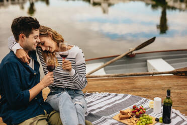 Romantic couple on a date sitting beside a lake with snacks and wine. Happy woman sitting with her boyfriend with her arm around his shoulders holding wine glasses. - JLPSF23592