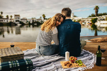 Rückansicht eines verliebten Paares, das Seite an Seite an einem See sitzt. Ein Paar, das an einem Tag mit Picknick und Getränken zusammensitzt und auf den See blickt, wobei die Frau zurückschaut. - JLPSF23595