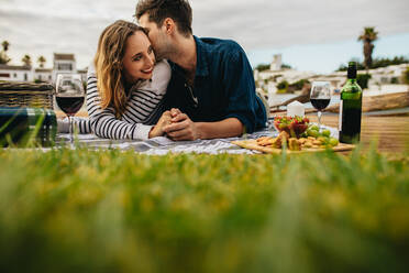 Couple on a date lying on ground drinking wine and snacks. Man kissing his girlfriend holding her hand at a date lying on a wooden dock with grass in the foreground. - JLPSF23597