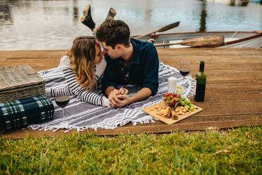 Verliebtes Paar, das auf einem hölzernen Steg neben einem See liegt, sich an den Händen hält und küsst. Romantisches Paar, das bei einem Date mit Getränken und Snacks Zeit miteinander verbringt, mit einem See im Hintergrund. - JLPSF23598