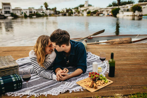 Romantisches Paar bei einem Date, das sich küsst und Zeit miteinander verbringt, während es in der Nähe eines Sees liegt. Ein Mann küsst seine Freundin, die auf einem hölzernen Steg in der Nähe eines Sees liegt und ihre Hand hält, mit Getränken und Essen an der Seite. - JLPSF23599