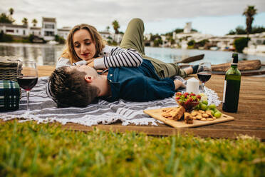 Ein Mann liegt auf dem Steg in der Nähe des Sees mit seiner Freundin, die ihre Hände auf seiner Brust ruhen lässt. Romantisches Paar bei einem Date in der Nähe eines Sees, das Essen und Getränke genießt. - JLPSF23600