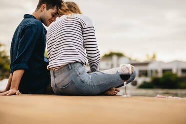 Rückansicht eines jungen Paares, das am Ufer eines Sees sitzt und sich verliebt die Köpfe berührt. Verliebtes Paar bei einem Rendezvous im Freien an einem See mit einem Glas Wein. - JLPSF23604