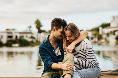 Junger Mann und Frau in einer romantischen Stimmung sitzen zusammen in der Nähe eines Sees. Junger Mann sitzt mit seiner Freundin in der Nähe eines Sees mit seinem Arm um ihren Hals. - JLPSF23606