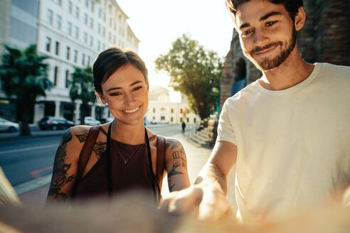 Tourist couple using a map to explore the city. Smiling man and woman standing by a street looking at map with sun in the background. - JLPSF23612