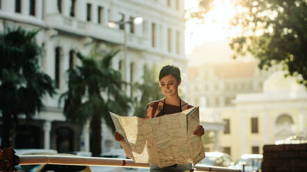 Lächelnde Frau, die auf der Straße steht und auf die Karte schaut, mit den Gebäuden der Stadt im Hintergrund. - JLPSF23613
