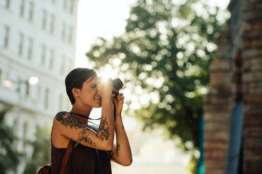 Frau Reisende fotografiert mit ihrer Kamera mit Sonne im Hintergrund. Tourist mit Tattoo auf der Hand, der durch die Stadt geht und fotografiert. - JLPSF23615