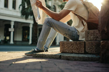 Traveler sitting on the pavement looking at a map with sun in the background. Tourist exploring the city with the help of a map. - JLPSF23616