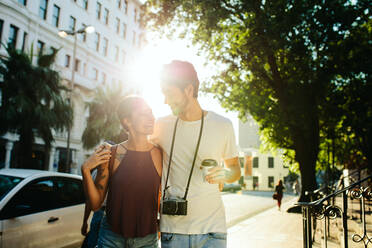 Glückliches Entdeckerpaar, das auf der Straße spazieren geht und eine Kaffeetasse hält, mit der Sonne im Hintergrund. Ein Mann trägt eine Kamera und hält eine Kaffeetasse, während er mit einer Frau spazieren geht und den Arm um ihre Schultern legt. - JLPSF23618