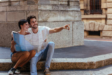 Touristisches Paar, das eine Karte benutzt, um den Weg zu seinem Ziel zu finden. Ein Mann und eine Frau, die auf dem Bürgersteig sitzen und eine Karte halten, wobei der Mann auf ein Objekt zeigt. - JLPSF23620