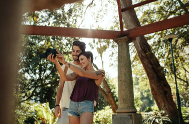 Glückliches Entdeckerpaar macht ein Selfie in einem Garten mit Sonne im Hintergrund. Glücklich gestimmtes Touristenpaar erkundet die Stadt. - JLPSF23623