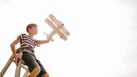 Junge sitzt auf einer Leiter mit einem Spielzeugflugzeug im hellen Sonnenlicht und stellt sich vor, er sei Pilot und fliege ein Spitzenflugzeug. - JLPSF23628