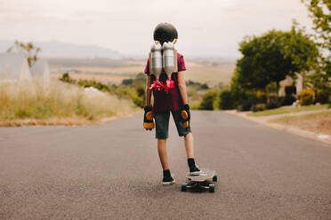 Rückansicht eines Jungen mit Helm und Spielzeug-Jetpack, der mit seinem Skateboard steht und die Straße hinunterschaut. Jetpack-Junge mit Skateboard - JLPSF23636