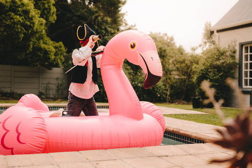Kleiner Junge als Pirat verkleidet, der auf einer aufblasbaren Flamingo-Matratze steht und durch sein Fernrohr in den Pool schaut. Junge, der vorgibt, ein Pirat zu sein, der im Swimmingpool spielt. - JLPSF23639