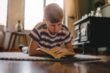 Kind liegt auf dem Boden und liest ein Buch. Junge entspannt sich auf einem Teppich und liest ein Buch zu Hause. - JLPSF23645