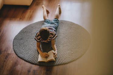 Draufsicht auf einen Jungen, der auf dem Boden liegt und ein Buch liest. Junge liest ein Buch zu Hause. - JLPSF23646
