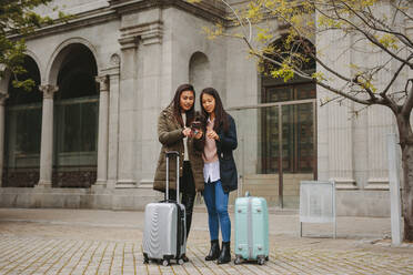 Zwei asiatische Touristinnen, die auf ihr Handy schauen, um zu navigieren. Weibliche Touristen, die mit Gepäckstücken auf der Straße stehen und nach einer Wegbeschreibung im Handy suchen. - JLPSF23651