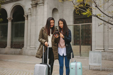 Touristinnen stehen auf der Straße mit Gepäckstücken und suchen nach einer Wegbeschreibung im Handy. Zwei lächelnde asiatische Touristen schauen auf ihr Handy. - JLPSF23652
