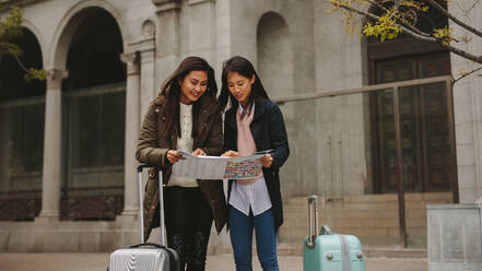 Zwei asiatische Touristinnen, die auf eine Karte schauen, um zu navigieren. Weibliche Touristen, die mit Gepäckstücken auf der Straße stehen und in einem Stadtplan nach dem Weg suchen. - JLPSF23654
