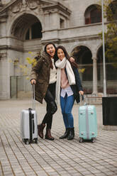 Two asian tourists exploring the city and having fun. Smiling female tourists standing together on street holding their luggage. - JLPSF23657