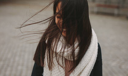 Close up of a woman in winter clothes standing outdoors with her hair covering her face. Smiling woman standing on street with hair flying on face. - JLPSF23662