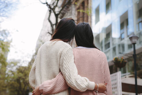 Frauen in Winterkleidung stehen zusammen und halten sich gegenseitig. Rückansicht von zwei Frauen in Pullovern, die im Freien stehen und sich umarmen. - JLPSF23665