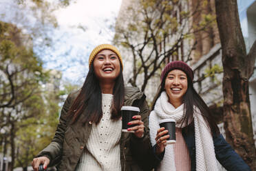 Lächelnde Frauen in Winterkleidung, die auf der Straße spazieren gehen und Kaffee trinken. Asiatische Touristinnen, die an einem Wintermorgen durch die Stadt laufen. - JLPSF23666