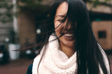 Lächelnde Frau auf der Straße mit fliegenden Haaren im Gesicht. Nahaufnahme einer asiatischen Frau in Winterkleidung, die im Freien steht und deren Haare ihr Gesicht verdecken. - JLPSF23669