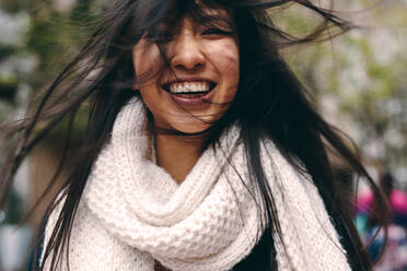 Close up of a woman standing outdoors with her hair covering her face. Smiling woman standing in street with her hair flying around. - JLPSF23670