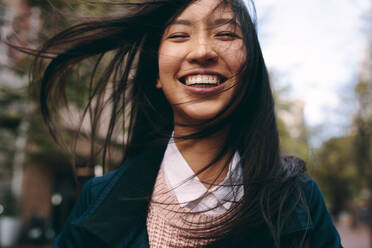 Porträt einer lächelnden asiatischen Frau, die im Freien steht. Lächelnde Frau, die auf der Straße steht und deren Haare herumfliegen. - JLPSF23671