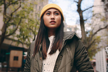 Close up of a woman in winter clothes standing outdoors. Asian woman standing in the street wearing a winter cap and looking away. - JLPSF23672