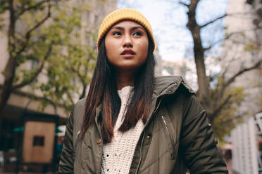 Nahaufnahme einer Frau in Winterkleidung, die im Freien steht. Asiatische Frau steht auf der Straße, trägt eine Wintermütze und schaut weg. - JLPSF23672